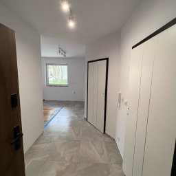 JS INVEST JAKUB DUNST - Wykończony korytarz z szarymi płytkami, białymi ścianami i drzwiami z czarnymi obramowaniami. Widoczne oświetlenie sufitowe i częściowo odsłonięta podłoga w pokoju.