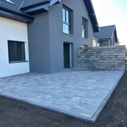 JS INVEST JAKUB DUNST - Nowoczesny taras z szarych płyt brukowych przy domu z szarą elewacją i ciemną stolarką okienną. Widoczny fragment muru z bloczków betonowych.