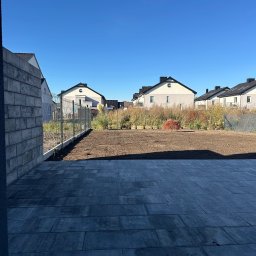 JS INVEST JAKUB DUNST - Ułożona szara kostka brukowa na tarasie, widok na przygotowany teren pod trawnik i ogrodzenie z siatki. W tle zabudowa szeregowa w słoneczny dzień.