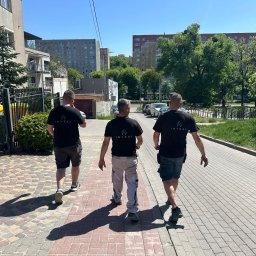 JS INVEST JAKUB DUNST - Trzech mężczyzn w czarnych koszulkach z logo idzie po brukowanej drodze w słoneczny dzień. W tle bloki mieszkalne i zieleń.