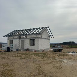 JS INVEST JAKUB DUNST - Budowa domu jednorodzinnego w stanie surowym otwartym. Widoczne ściany z bloczków, więźba dachowa, przyczepa i materiały budowlane. Pochmurne niebo w tle.