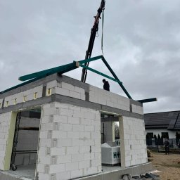 JS INVEST JAKUB DUNST - Budowa domu z białych bloczków H+H. Żuraw montuje drewniane elementy dachu. Widoczny pracownik na ścianie budynku i materiały budowlane.