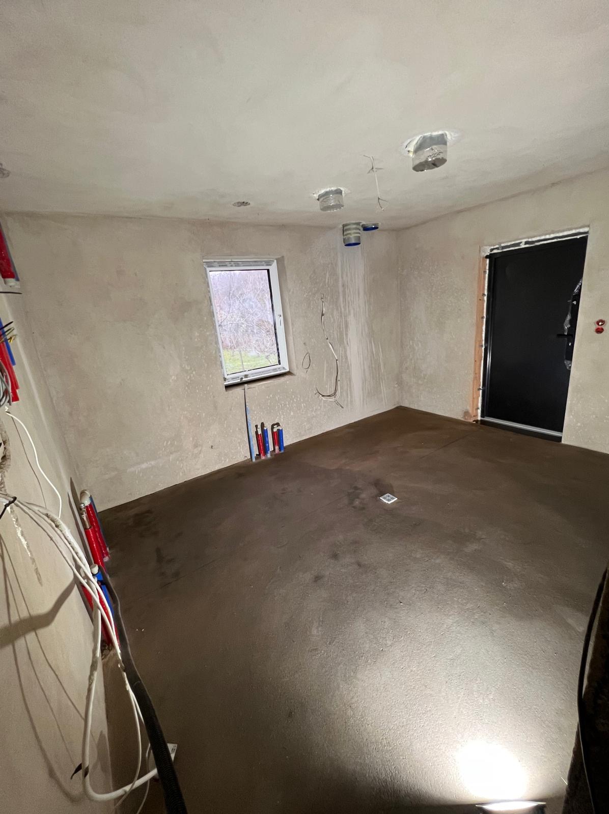 Wnętrze w trakcie prac: wylana posadzka, surowe ściany, instalacje elektryczne i hydrauliczne. Widoczne okno z widokiem na zewnątrz, czarne drzwi.