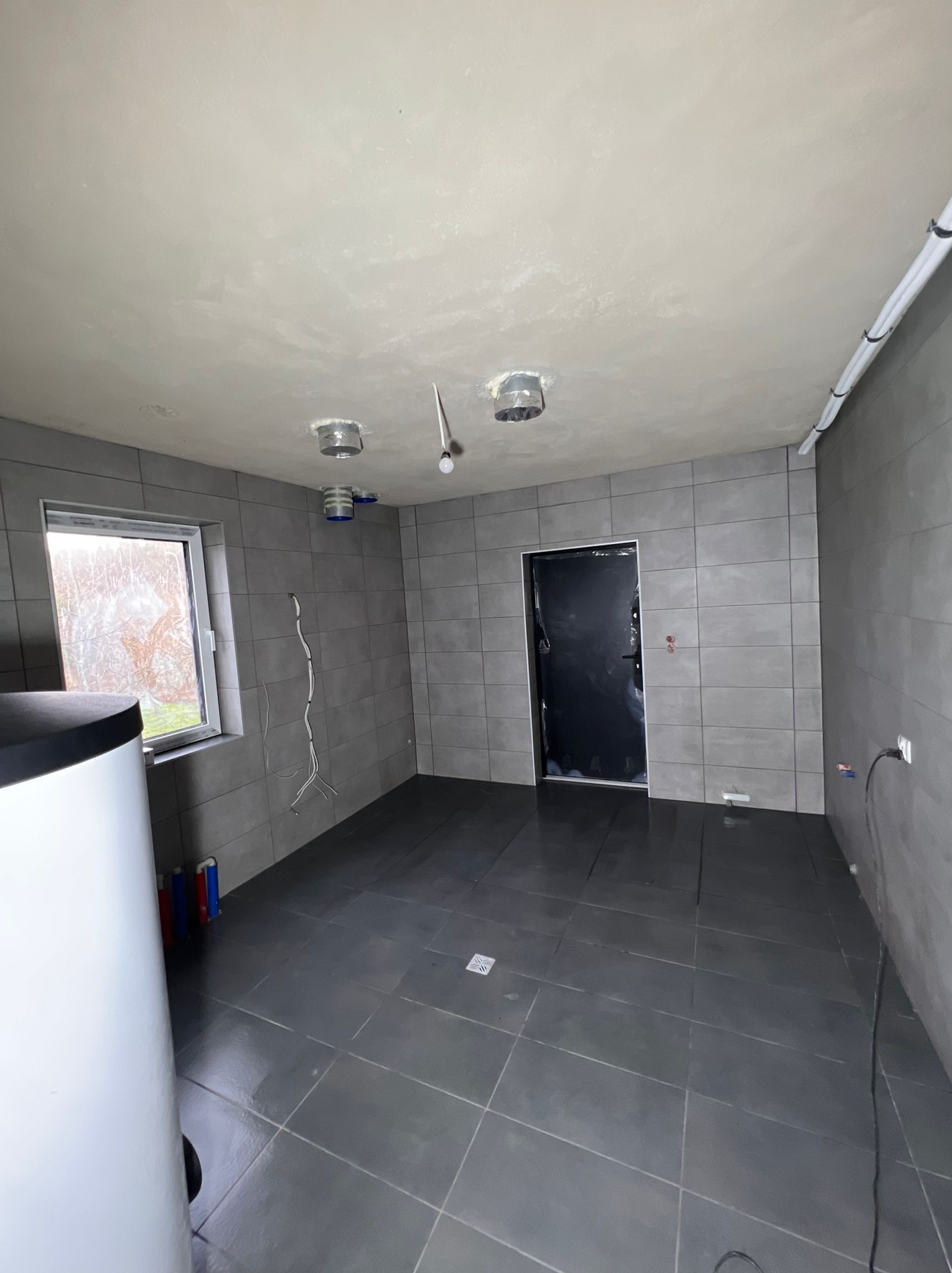 Szara łazienka w trakcie wykończenia: płytki na ścianach i podłodze, biały bojler, przygotowane punkty świetlne i instalacja elektryczna. Drzwi zabezpieczone folią.