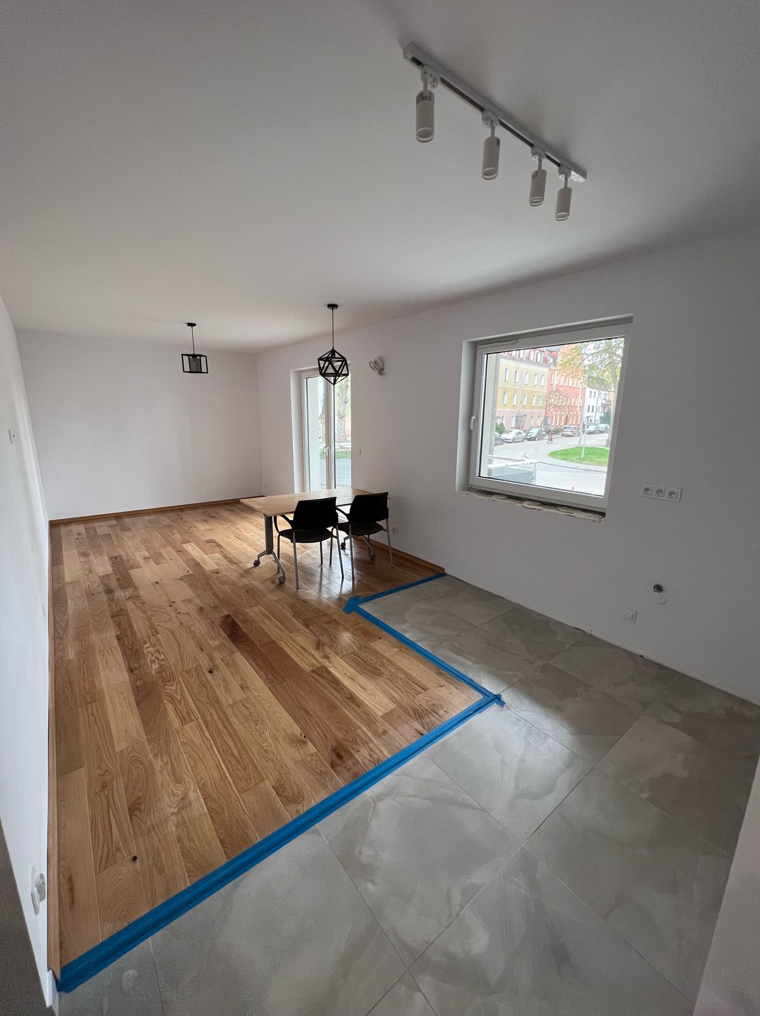 Jasne, przestronne wnętrze po częściowym remoncie: parkiet dębowy ułożony obok szarych płytek. Niebieska taśma oddziela powierzchnie. Widoczne lampy i okno z widokiem na miasto.