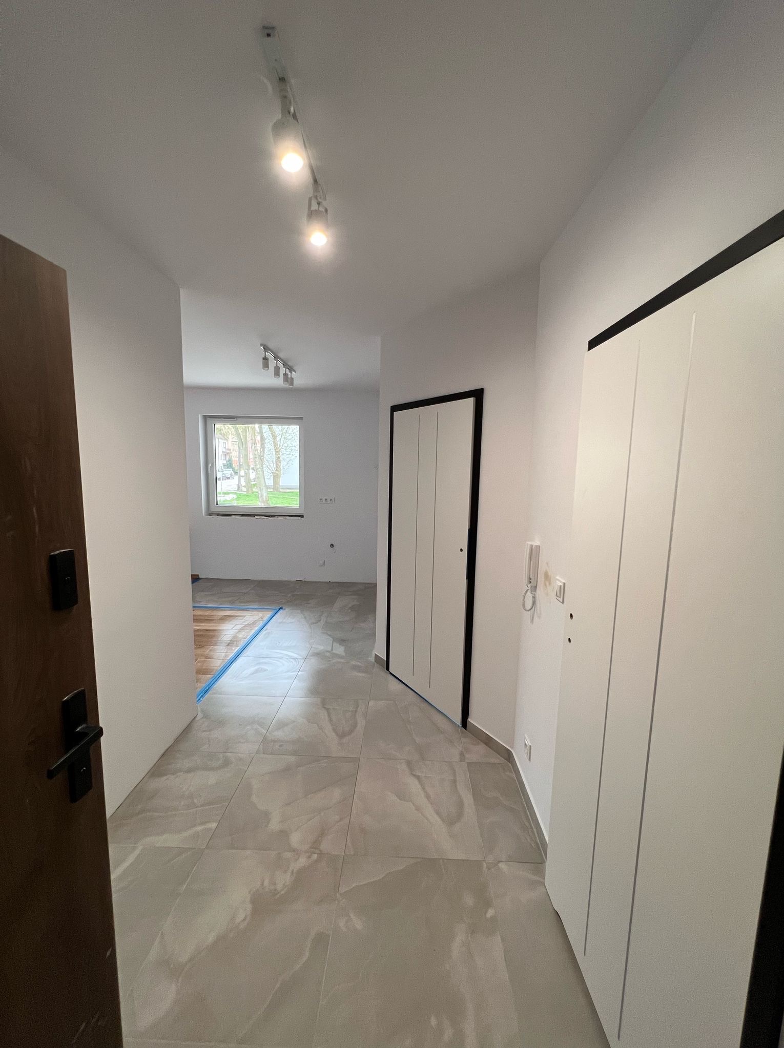 Wykończony korytarz z szarymi płytkami, białymi ścianami i drzwiami z czarnymi obramowaniami. Widoczne oświetlenie sufitowe i częściowo odsłonięta podłoga w pokoju.