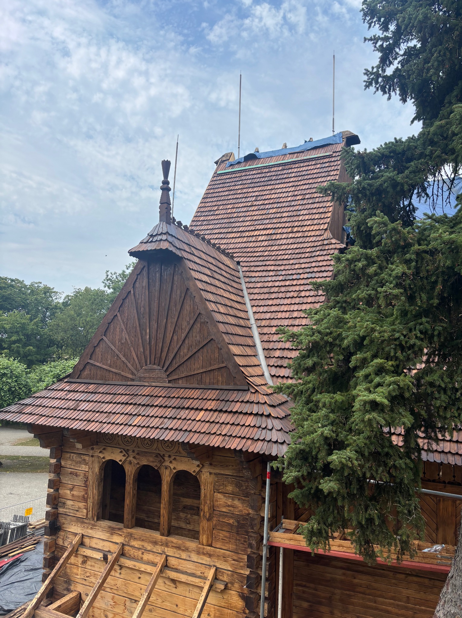 Drewniany budynek z czerwonym dachem z dachówki karpiówki, częściowo pod plandeką, z dekoracyjnymi elementami i rusztowaniem. Widok z góry, drzewa w tle.