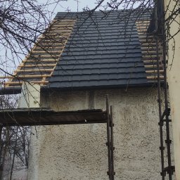 FIRMA REMONTOWO-BUDOWLANA SUDETY SPÓŁKA Z OGRANICZONĄ ODPOWIEDZIALNOŚCIĄ - Dom w trakcie prac dekarskich: częściowo ułożona grafitowa dachówka, widoczne drewniane łaty, rusztowanie i gałęzie drzew na pierwszym planie.