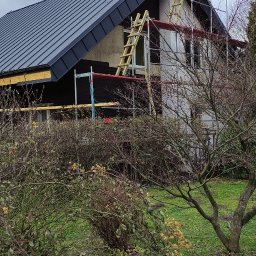 FIRMA REMONTOWO-BUDOWLANA SUDETY SPÓŁKA Z OGRANICZONĄ ODPOWIEDZIALNOŚCIĄ - Dom w trakcie prac dekarskich: nowy, grafitowy dach z blachy na ukończeniu, widoczne rusztowania i drabiny, częściowo ocieplona elewacja, w tle pochmurne niebo.