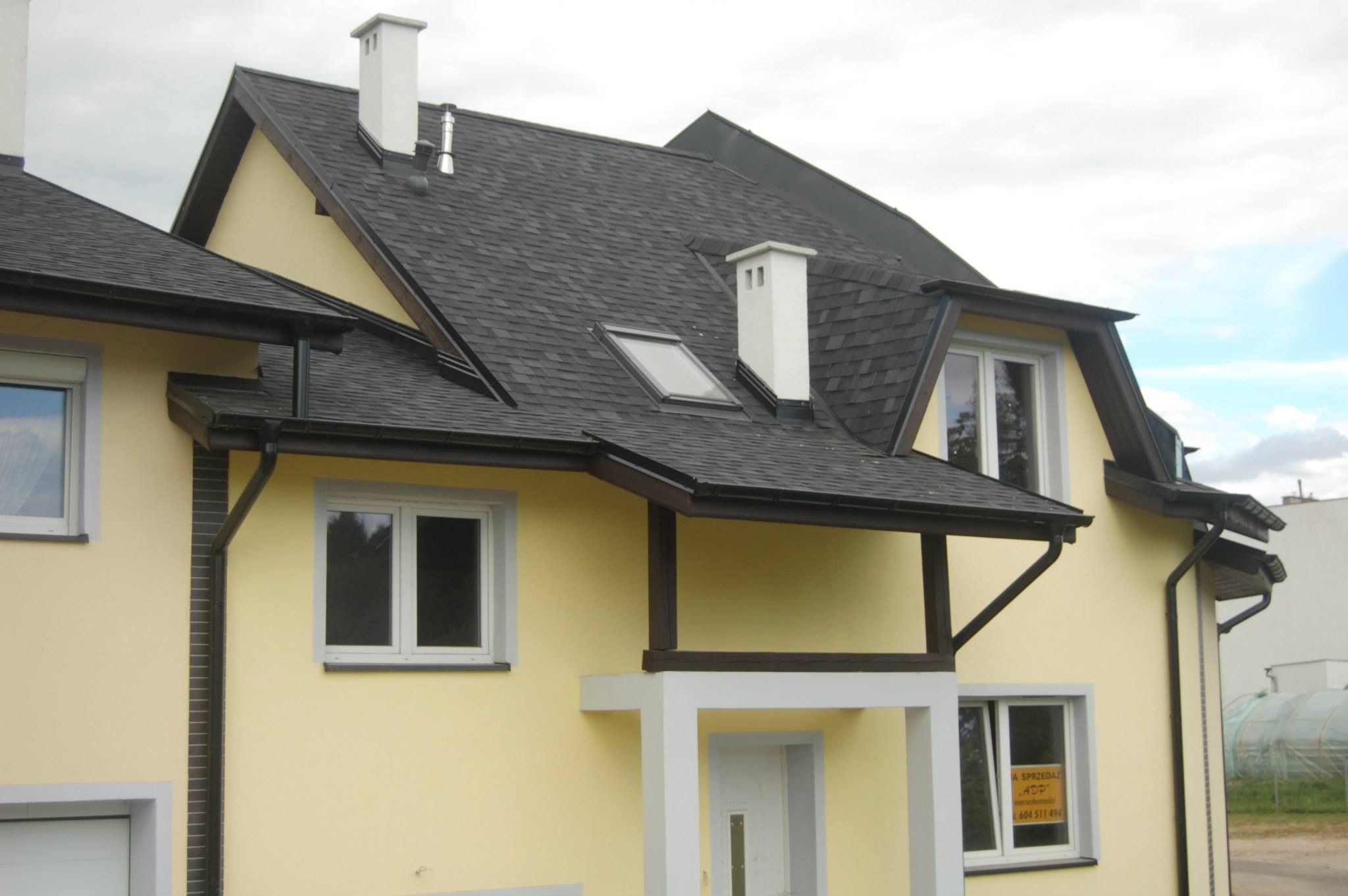 Żółty dom jednorodzinny z czarnym dachem bitumicznym, białymi kominami i oknem dachowym; widoczny fragment elewacji z rynnami i oknem z ogłoszeniem 'A SPRZEDAŻ'.