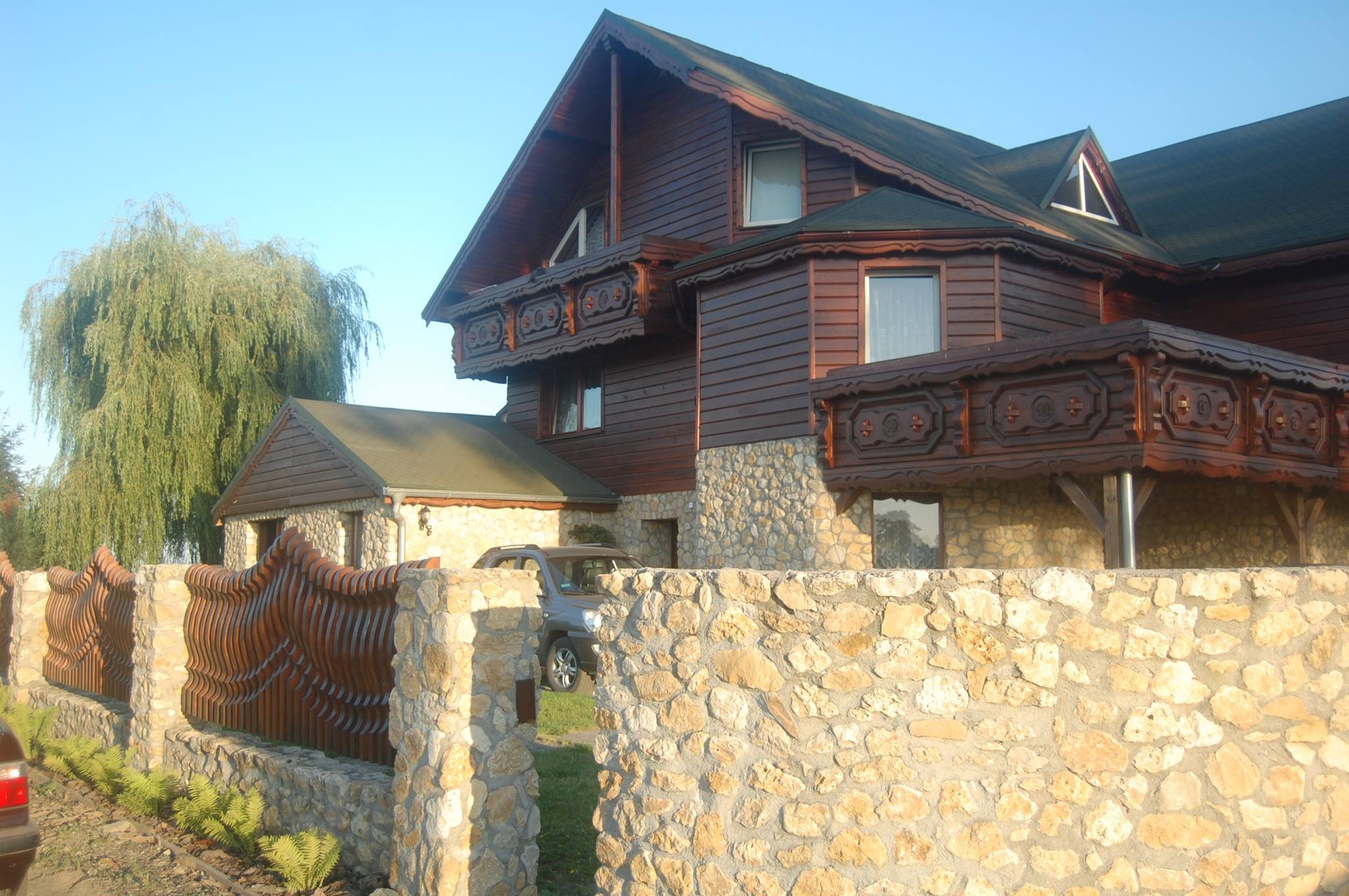 Dom szkieletowy z drewnianą elewacją i zdobionymi balkonami w Prudniku, otoczony kamiennym murem i ozdobnym drewnianym płotem.