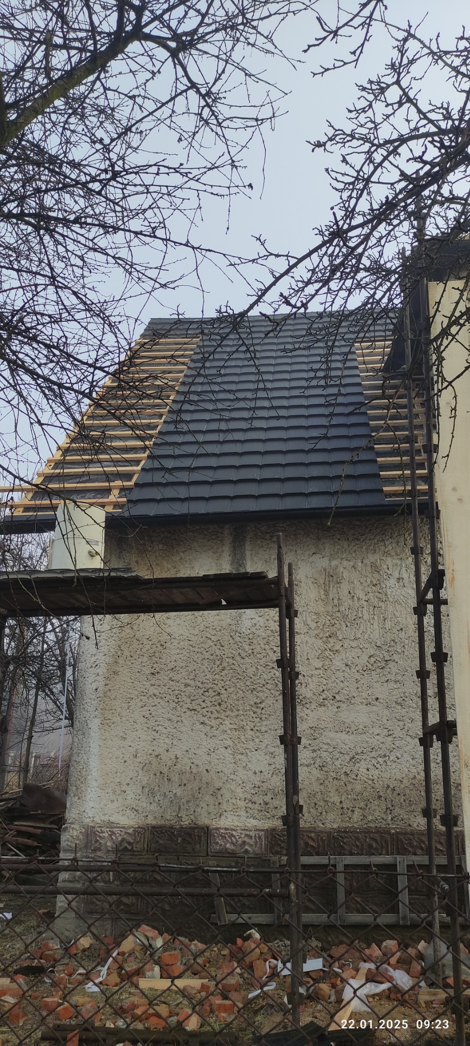 Dom w trakcie prac dekarskich: częściowo ułożona grafitowa dachówka, widoczne drewniane łaty, rusztowanie i gałęzie drzew na pierwszym planie.