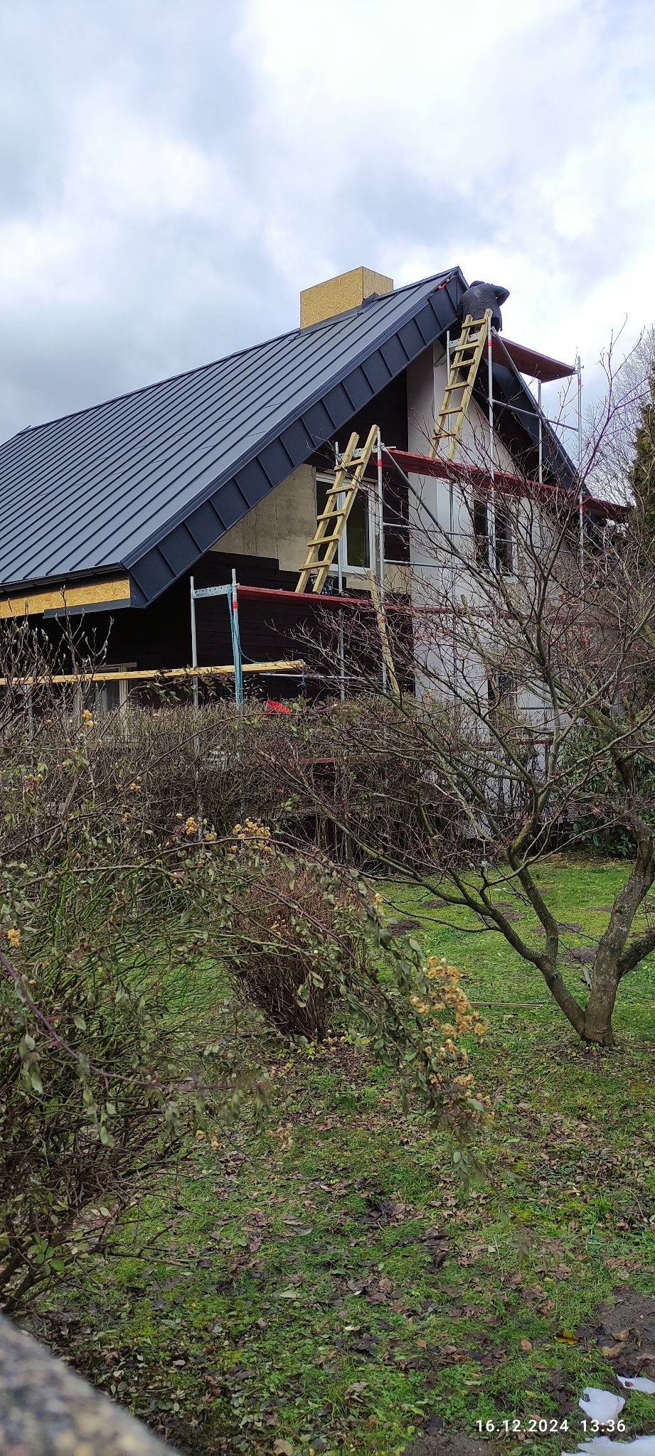 Dom w trakcie prac dekarskich: nowy, grafitowy dach z blachy na ukończeniu, widoczne rusztowania i drabiny, częściowo ocieplona elewacja, w tle pochmurne niebo.