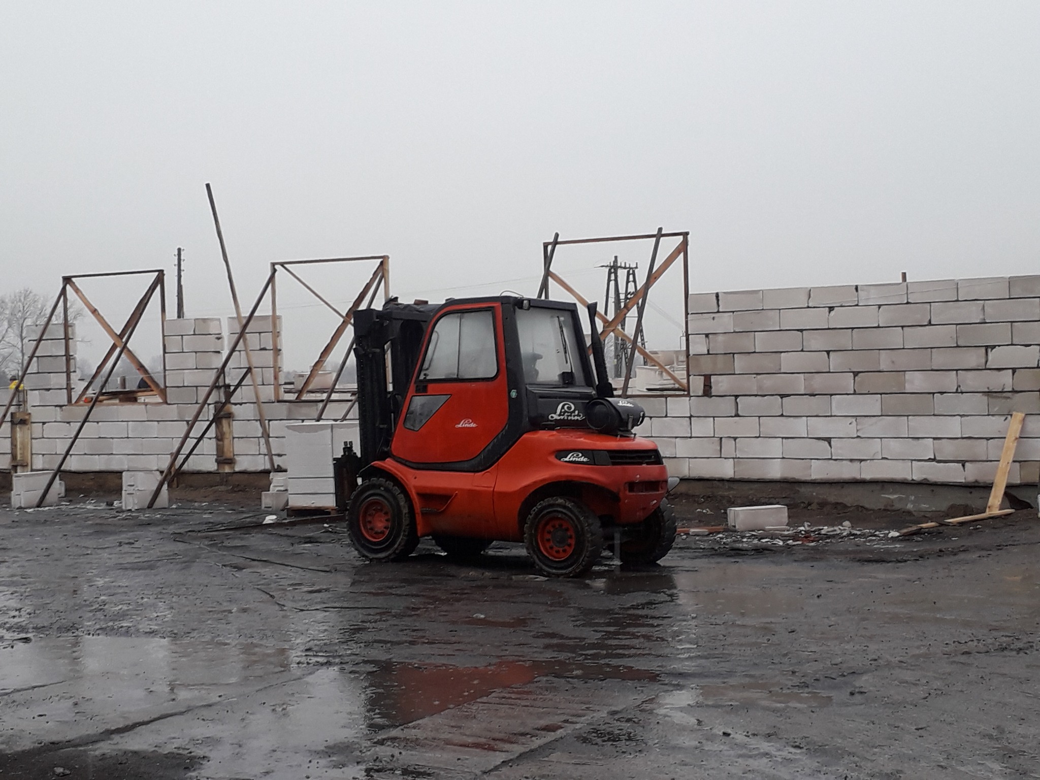 Pomarańczowy wózek widłowy Linde na mokrym placu budowy, w tle ściany z białych bloczków i drewniane konstrukcje szkieletowe.