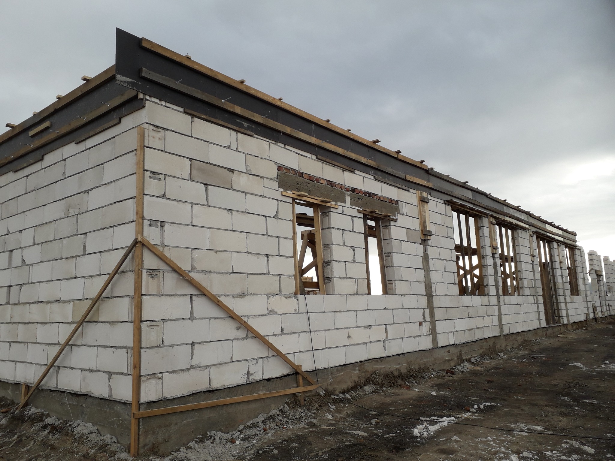 Surowy stan budowy domu z białych bloczków, widoczne drewniane elementy konstrukcyjne dachu i szalunki okienne, pochmurne niebo w tle.