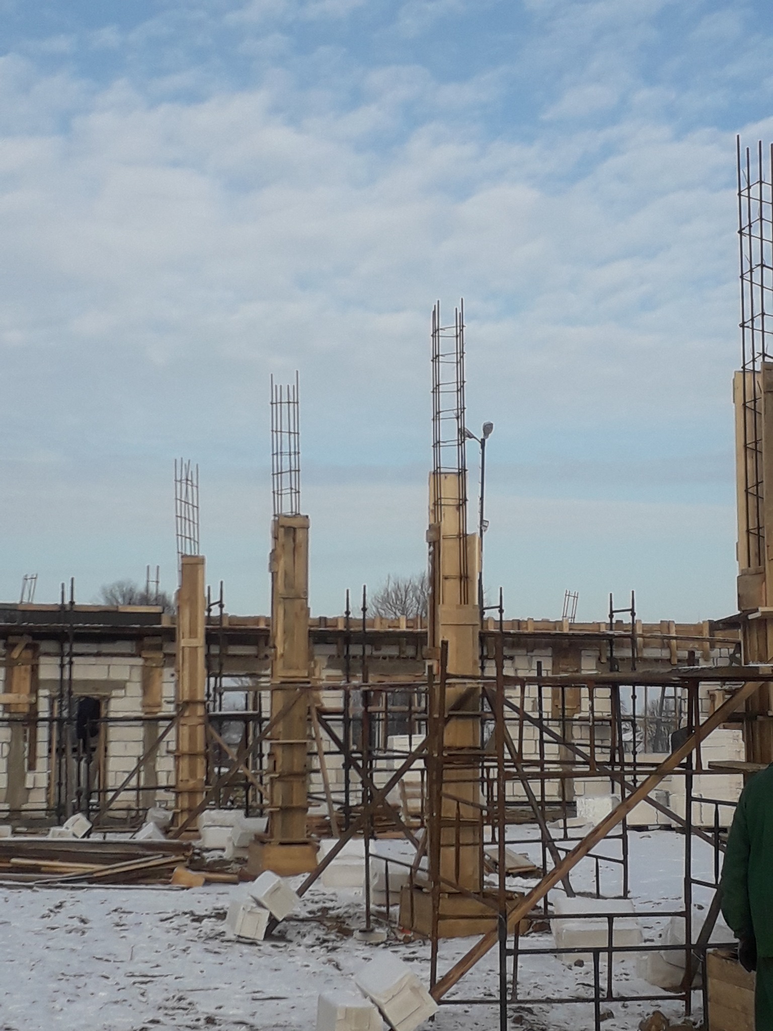 Budowa domu szkieletowego zimą: widoczne szkieletowe kolumny zbrojeniowe w drewnianych szalunkach, rusztowania i śnieg na placu budowy pod pochmurnym niebem.
