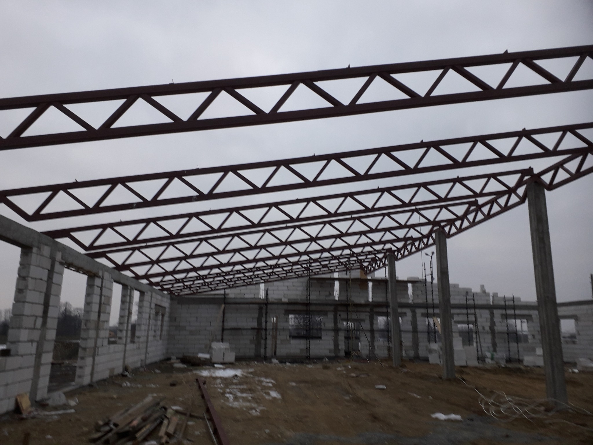 Budowa domu w stanie surowym otwartym: widoczne ściany z bloczków, stalowa konstrukcja dachu z elementami trójkątnymi, ziemia na podłodze, pochmurne niebo w tle.