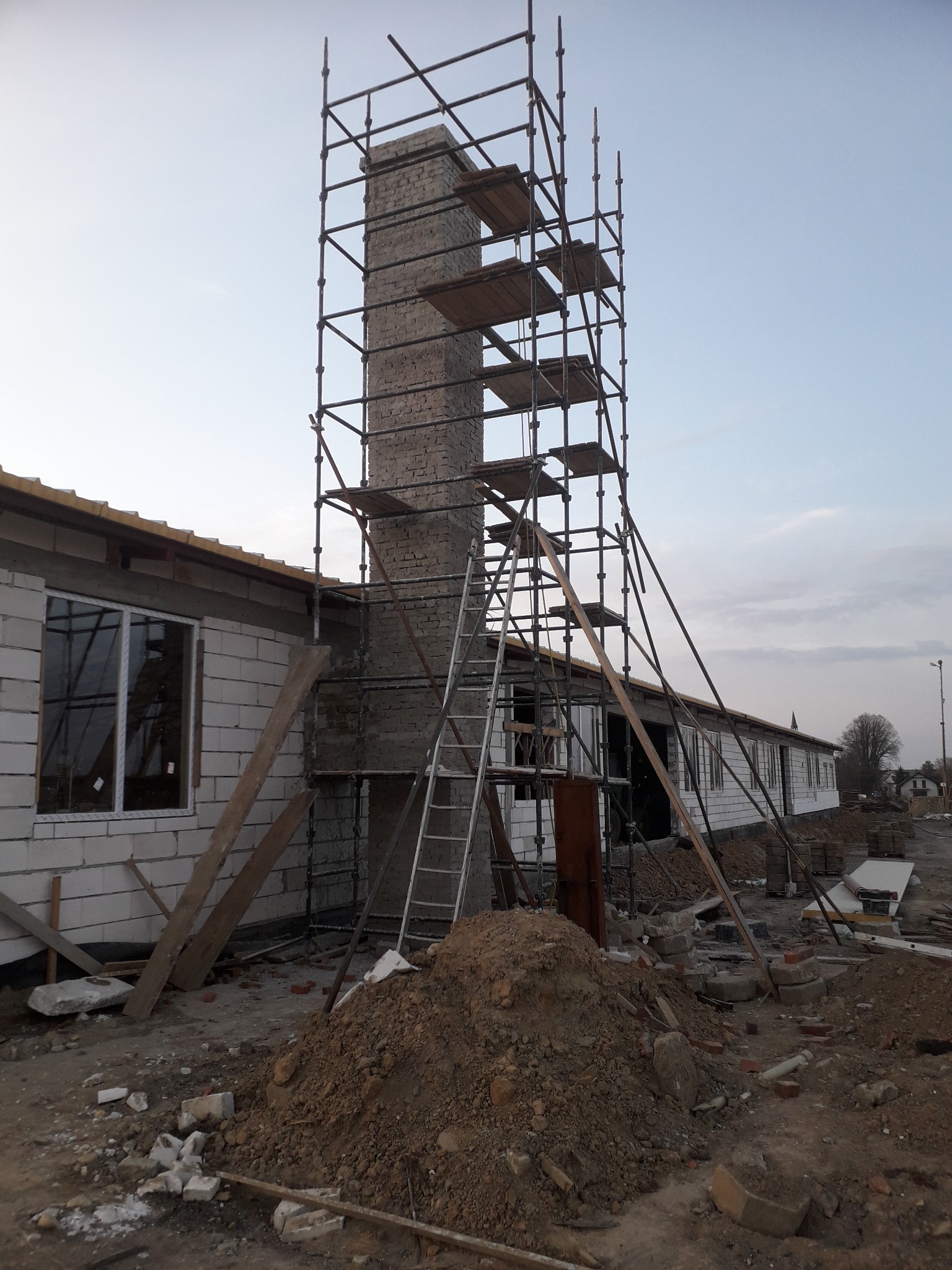 Budowa domu z pustaków: wznoszenie komina z rusztowaniem, widoczne elementy konstrukcyjne i sterta ziemi.