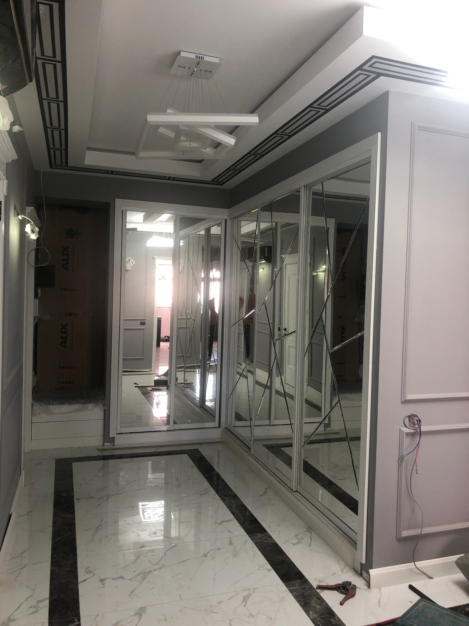 Nowoczesne wnętrze: białe ściany z dekoracyjnymi panelami, lustrzane drzwi przesuwne szafy wnękowej z geometrycznymi zdobieniami, marmurowa podłoga z czarnymi pasami, designerska lampa sufitowa.