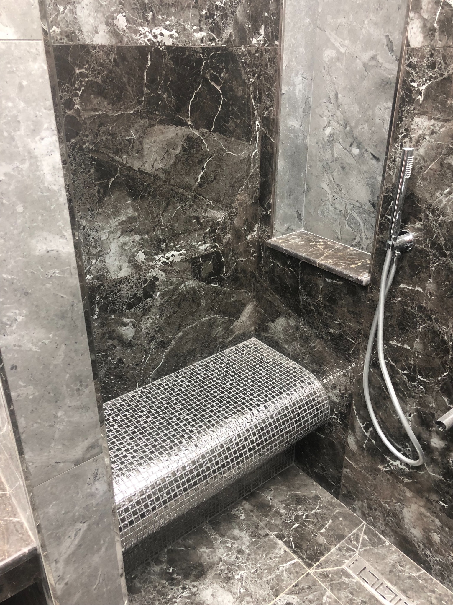 Kabina prysznicowa wykończona ciemnym marmurem z mozaikowym siedziskiem i półką wnękową, widoczna słuchawka prysznicowa.
