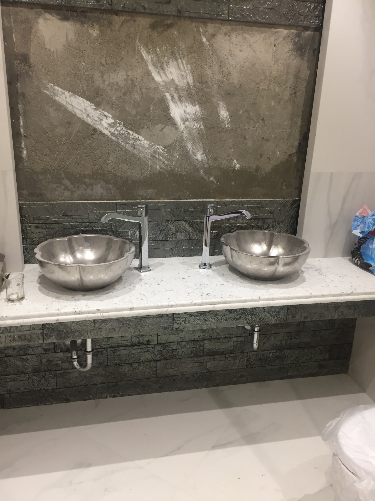 Dwie metalowe umywalki w kształcie kwiatów z chromowanymi bateriami na blacie z szarej mozaiki, w tle ściana w trakcie wykończenia z widocznymi śladami zaprawy.