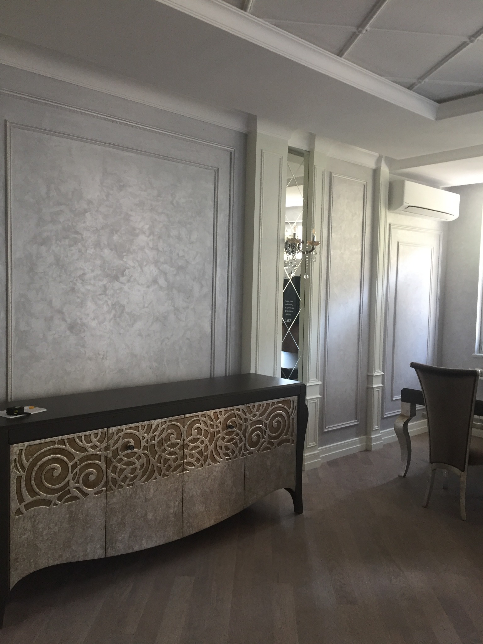 Eleganckie wnętrze jadalni z dekoracyjną ścianą w odcieniach szarości, sztukaterią i komodą z ornamentami, widok na stół i krzesła.