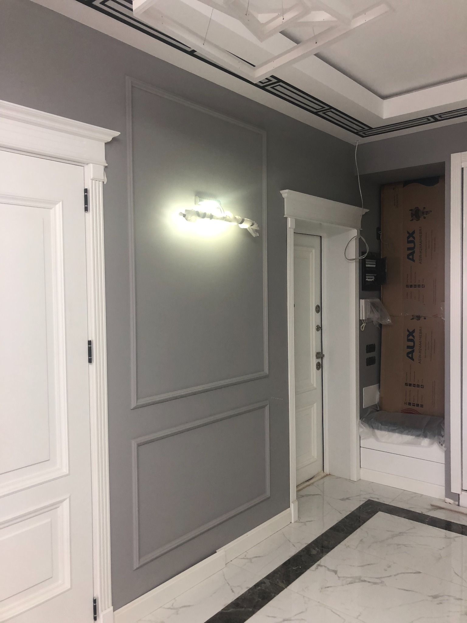 Szare ściany z dekoracyjnymi listwami i oświetleniem, białe drzwi z ozdobnymi framugami, marmurowa podłoga w geometryczny wzór, widoczne kartony w głębi pomieszczenia.