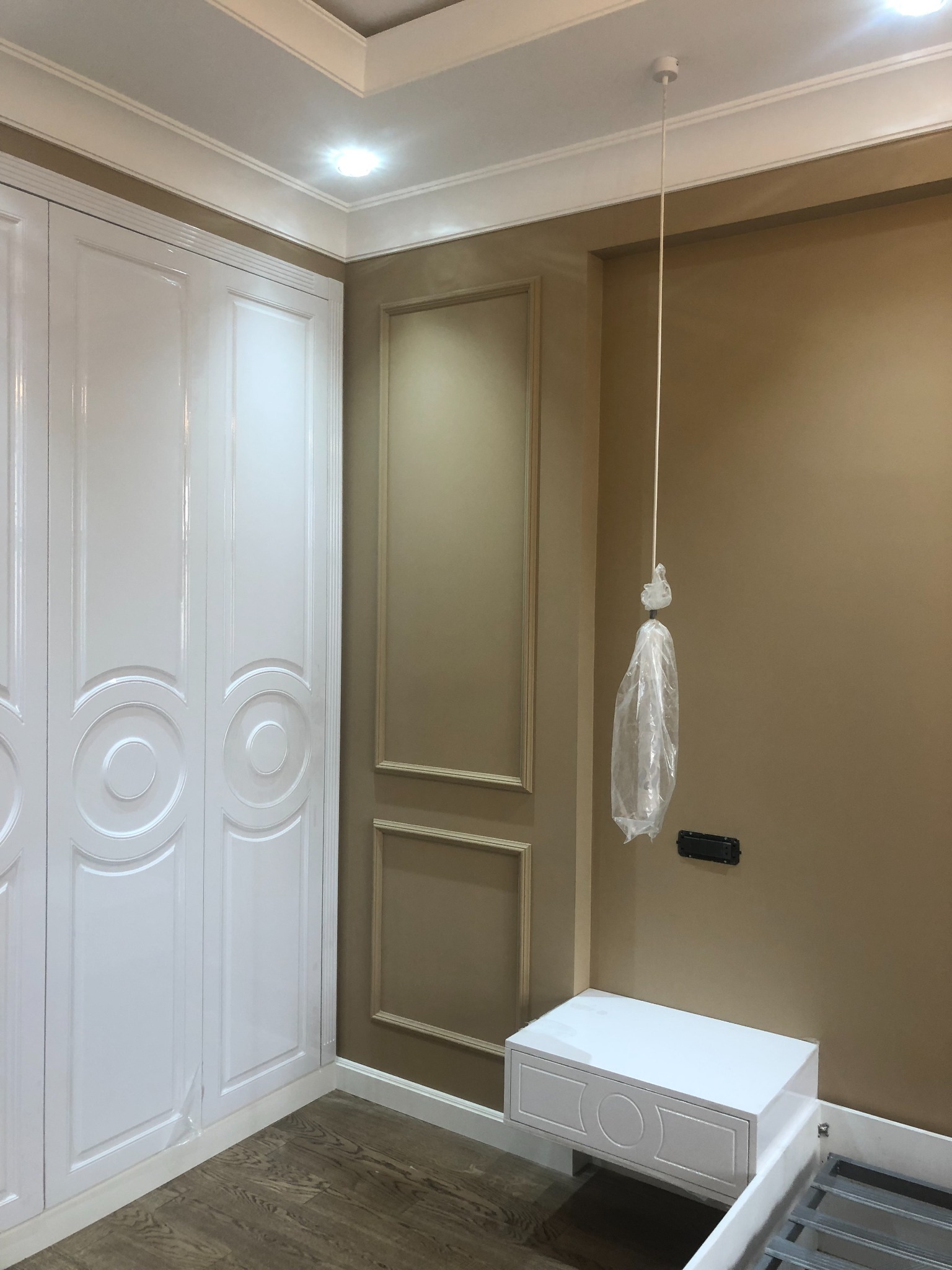 Pokój po remoncie: biała, lakierowana szafa z dekoracyjnymi okręgami, beżowa ściana z dekoracyjną ramą, biała szafka nocna i lampa wisząca zabezpieczona folią ochronną.