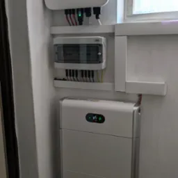 Biały falownik Huawei z podłączonymi kablami, skrzynka rozdzielcza i akumulator energii w pomieszczeniu z oknem.