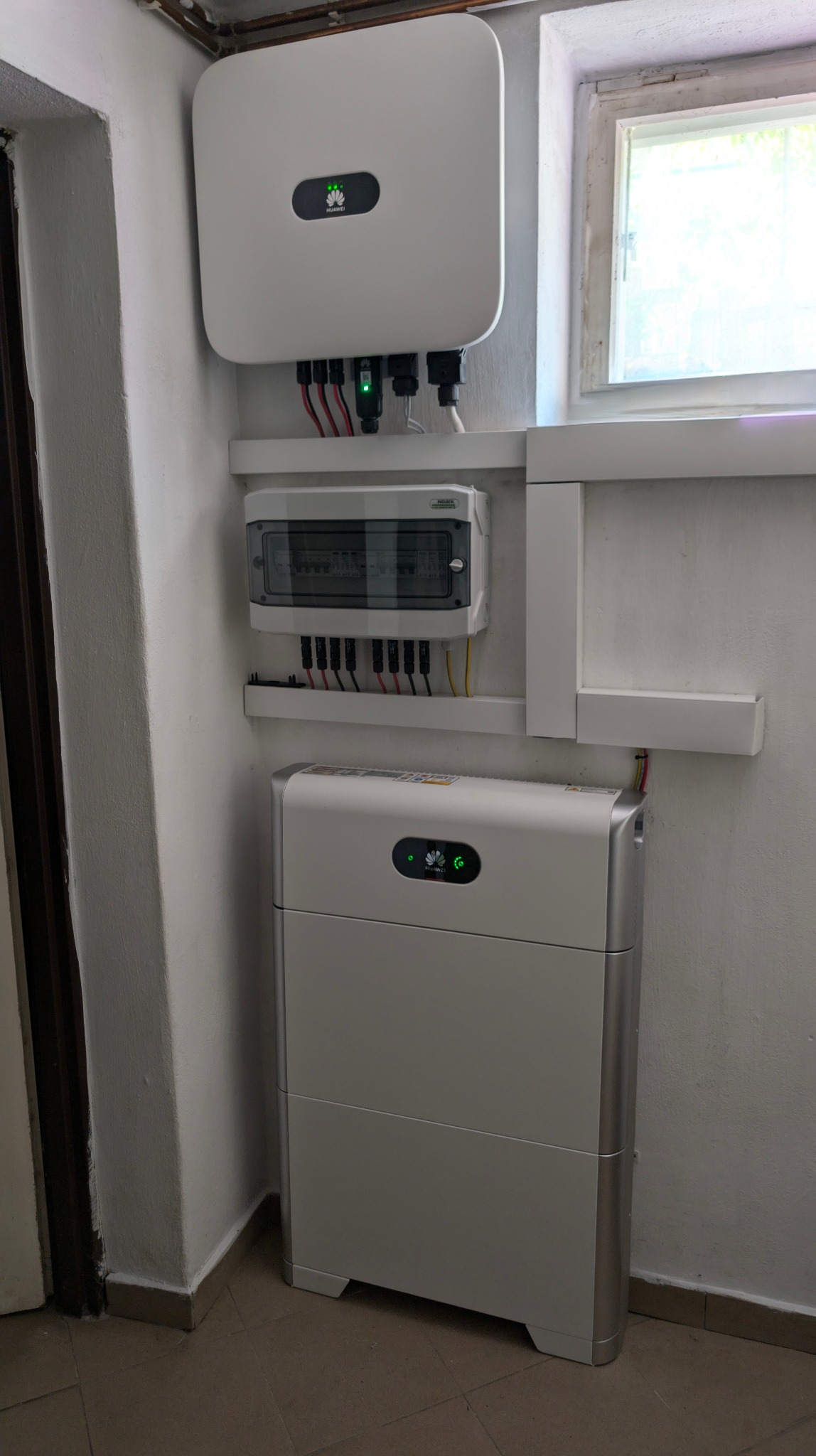 Biały falownik Huawei z podłączonymi kablami, skrzynka rozdzielcza i akumulator energii w pomieszczeniu z oknem.