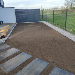 FIRMA OGRODNICZA GARDENOW SPÓŁKA CYWILNA - Przygotowany teren pod przyszły trawnik, obrzeża z jasnego grysu, płyty chodnikowe wkomponowane w ziemię, nowoczesne ogrodzenie panelowe w tle.