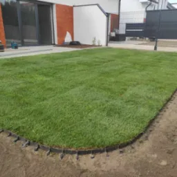 Świeżo ułożona darń z rolki w ogrodzie, widoczne czarne obrzeże trawnikowe z plastikowymi kołkami mocującymi, nowoczesny dom w tle.