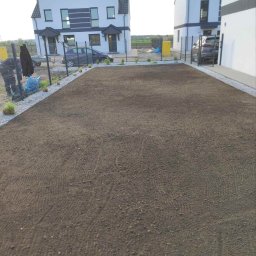 FIRMA OGRODNICZA GARDENOW SPÓŁKA CYWILNA - Przygotowany teren pod przyszły trawnik, widoczne świeżo posadzone małe rośliny wzdłuż ogrodzenia dwóch nowoczesnych domów.