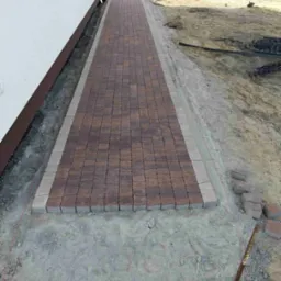 Świeżo ułożony chodnik z kostki brukowej w dwóch odcieniach brązu z szarym obramowaniem, widoczne narzędzia i teren po robotach ziemnych.