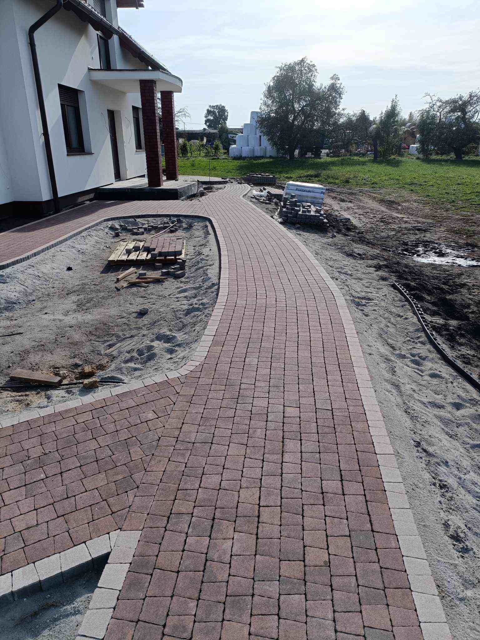 Układanie zakrzywionego chodnika z czerwonej kostki brukowej z szarą obwódką, widoczne przygotowane podłoże z piasku i palety z kostką.