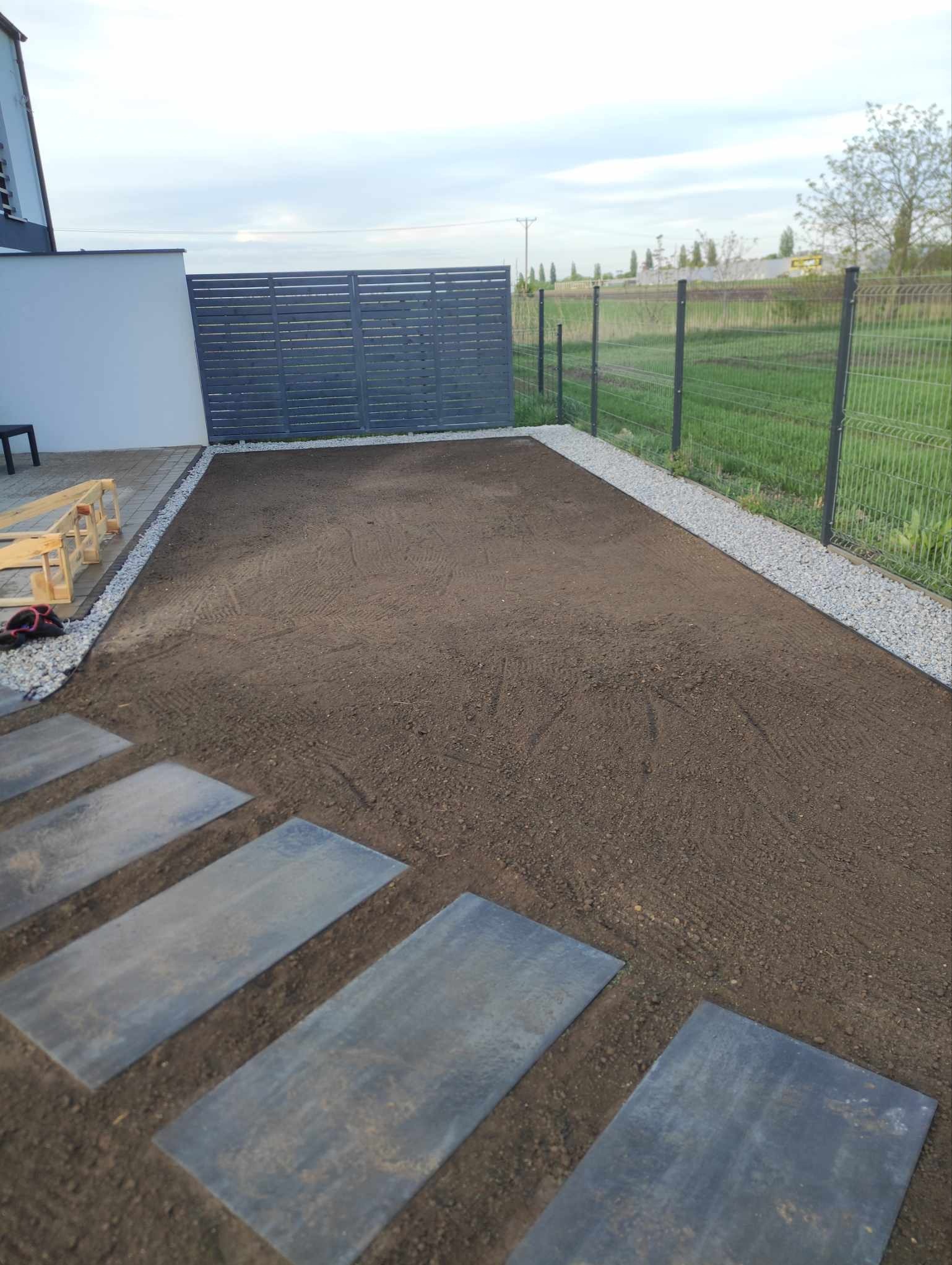 Przygotowany teren pod przyszły trawnik, obrzeża z jasnego grysu, płyty chodnikowe wkomponowane w ziemię, nowoczesne ogrodzenie panelowe w tle.