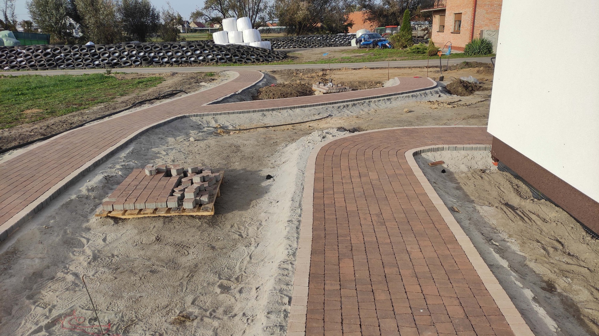 Kaskadowe ścieżki z brązowej kostki brukowej w trakcie układania, obrzeża z jasnych kostek, paleta z kostką w tle, opony ułożone w stos w oddali.