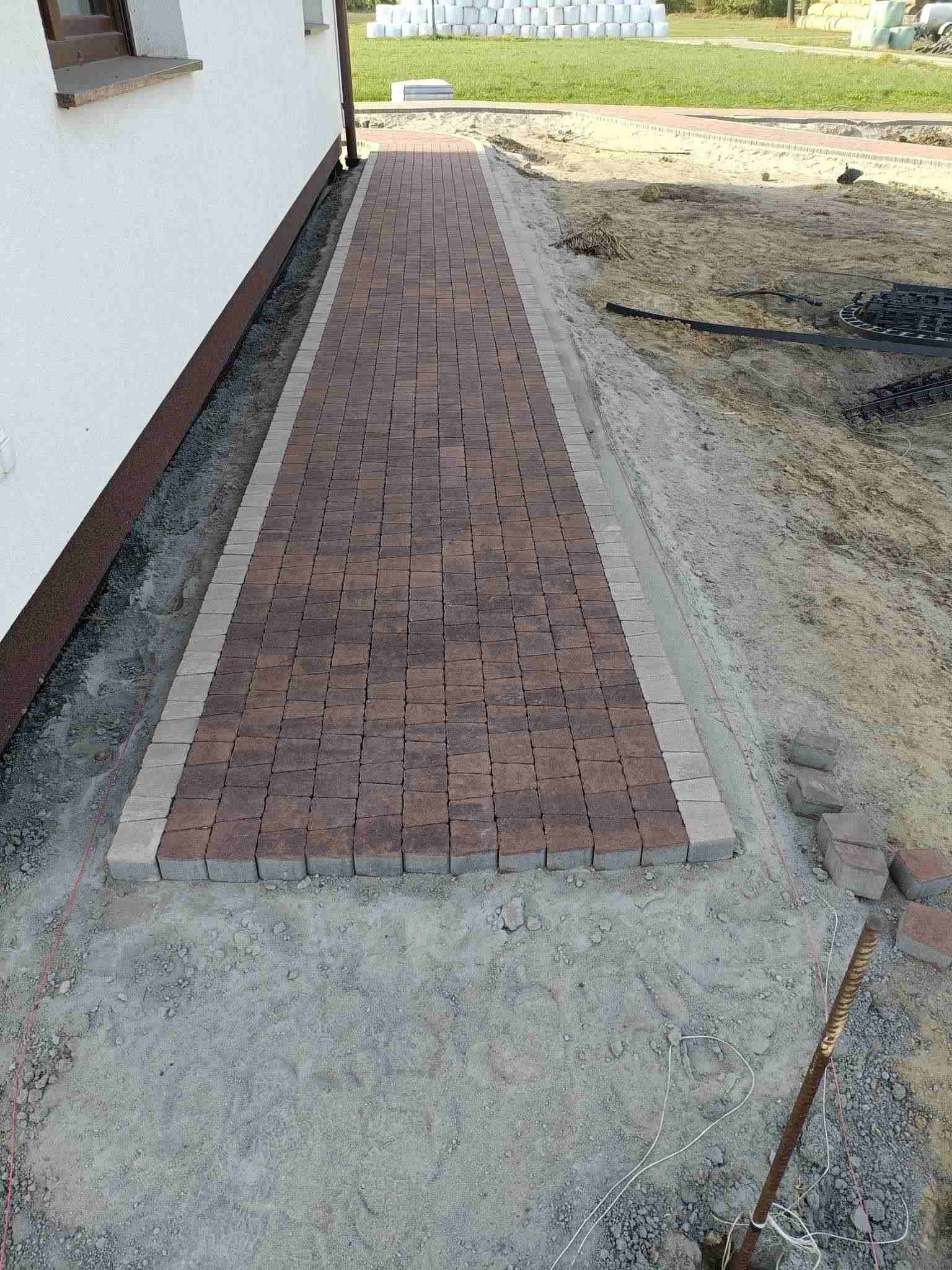 Świeżo ułożony chodnik z kostki brukowej w dwóch odcieniach brązu z szarym obramowaniem, widoczne narzędzia i teren po robotach ziemnych.