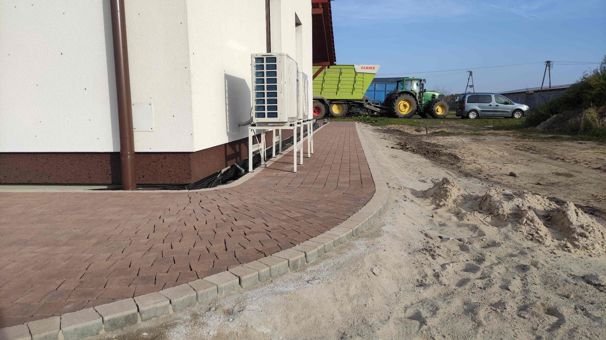 Nowo ułożona, brązowa kostka brukowa z szarą obwódką, prowadząca wzdłuż ściany budynku z zamontowanymi klimatyzatorami, w tle traktor z przyczepą i zaparkowany samochód.