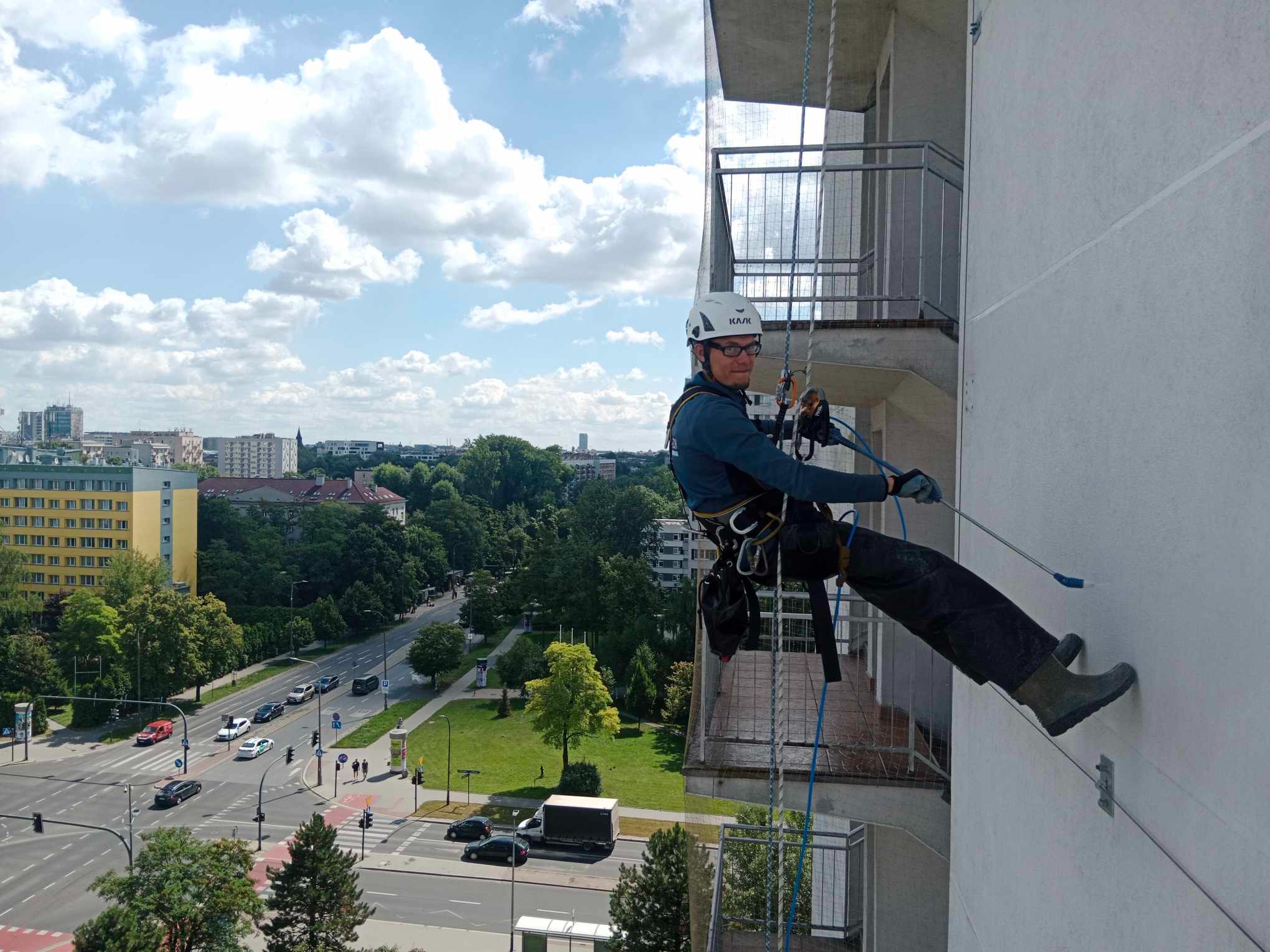 Pracownik w kasku i uprzęży, zawieszony na linie, czyści elewację budynku z wysokości, widok na miejski krajobraz z balkonu.