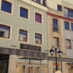 Elewacja budynku z szyldem 'Yasumi' nad wejściem, odbijająca się w szklanych drzwiach. Tablica informacyjna z logo Yasumi stoi przed wejściem. Budynek wielopiętrowy, z oknami w kolorze brązowym...