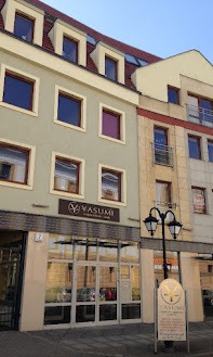 Elewacja budynku z szyldem 'Yasumi' nad wejściem, odbijająca się w szklanych drzwiach. Tablica informacyjna z logo Yasumi stoi przed wejściem. Budynek wielopiętrowy, z oknami w kolorze brązowym...