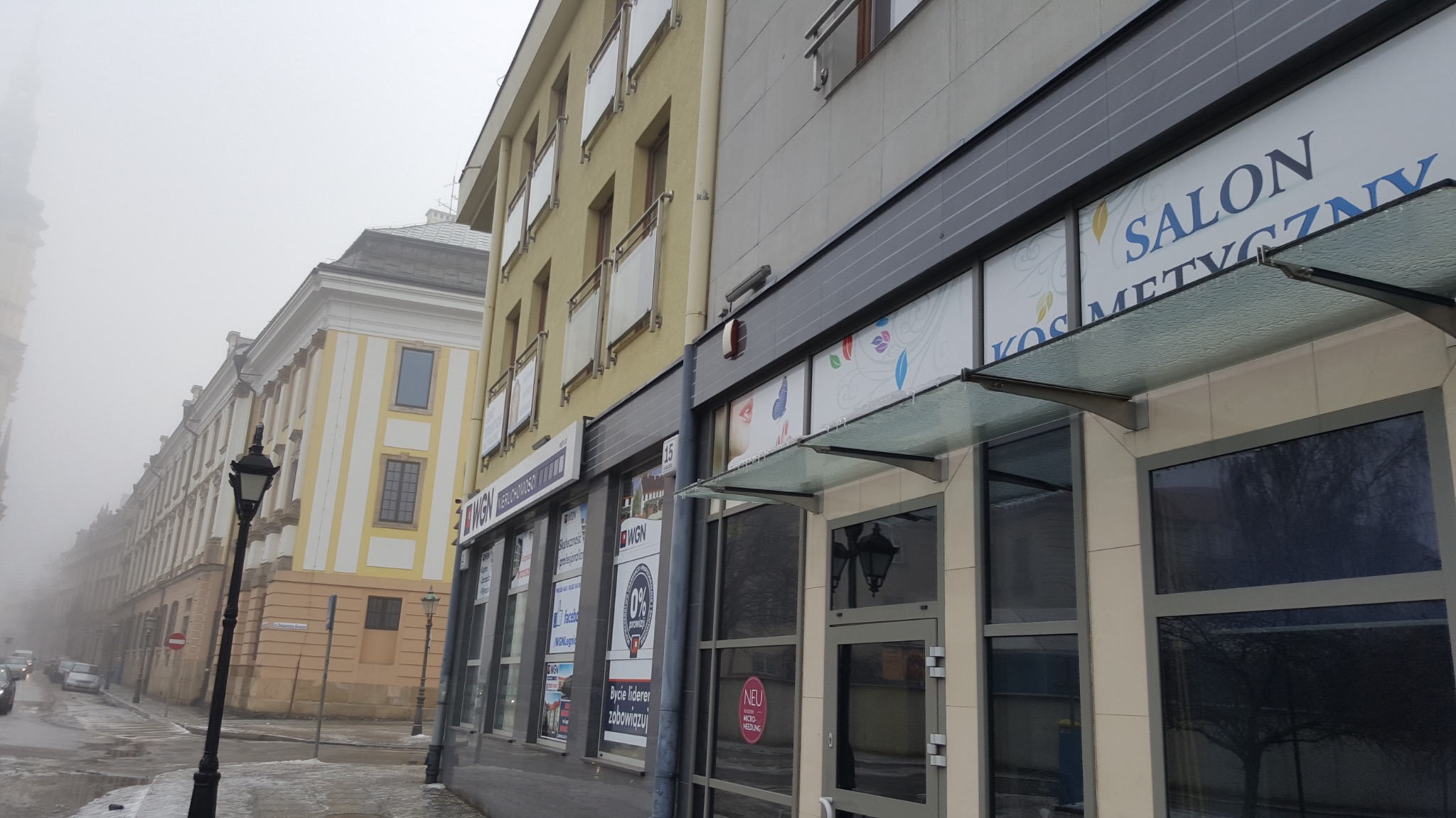 Fasada budynku w mglisty dzień, widoczny salon kosmetyczny z przeszklonym wejściem i szyldem 'Salon Kosmetyczny', obok biura nieruchomości z licznymi plakatami w oknach, latarnia uliczna w stylu...