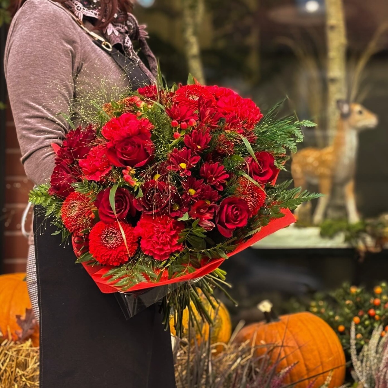 Osoba trzyma w dłoniach duży, okrągły bukiet intensywnie czerwonych róż, chryzantem i innych kwiatów, zawinięty w czerwony papier. W tle, za osobą, widoczne są dynie, snopki siana i dekoracja...