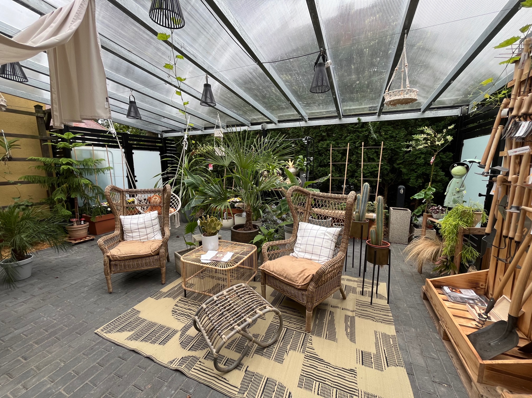 Zadaszona przestrzeń zewnętrzna z wiklinowymi fotelami, stolikiem i pufem na wzorzystym dywanie, otoczona różnorodną roślinnością w donicach i skrzyniach, z widocznymi narzędziami ogrodniczymi...