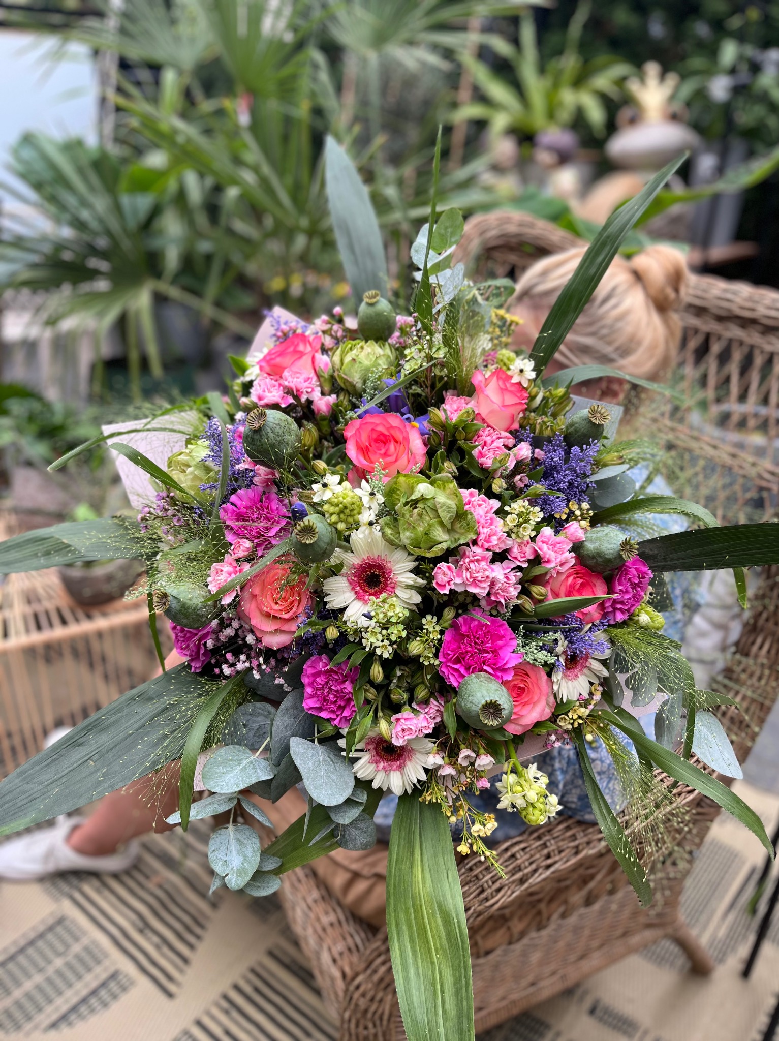 Bogaty bukiet różowych i białych róż, goździków, gerber i makówek, trzymany w wiklinowym fotelu na tle zieleni ogrodowej.