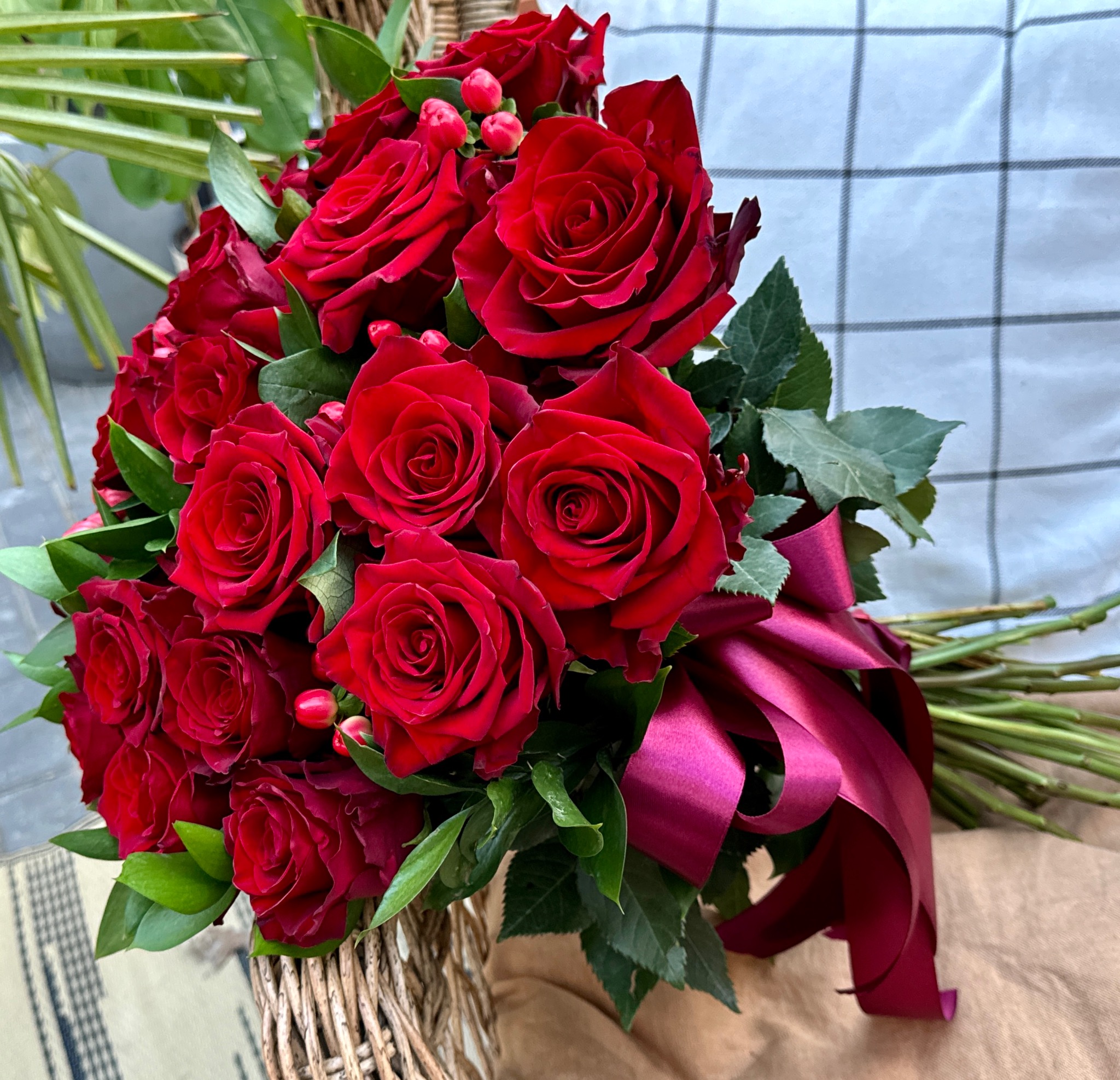 Bukiet czerwonych róż z bordową wstążką w wiklinowym koszu, na tle białej kraty.