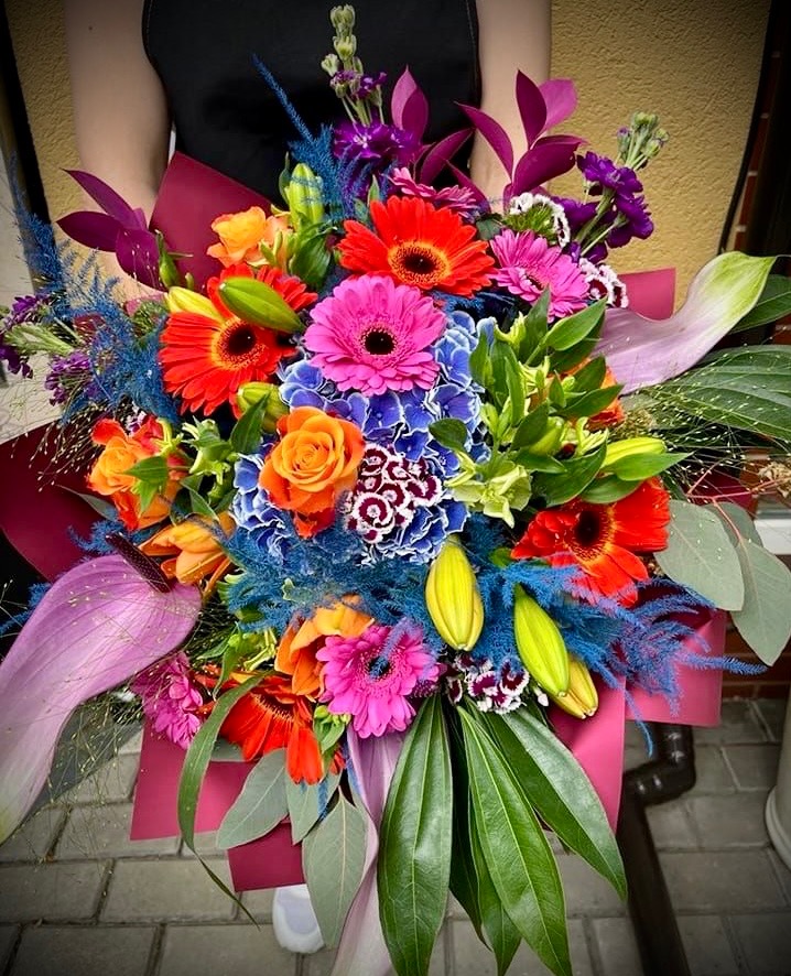 Barwny bukiet kwiatów z gerberami, różami, anturium i hortensjami, owinięty w bordowy papier, trzymany przez osobę w czarnej bluzce na tle beżowej ściany i szarej kostki brukowej.