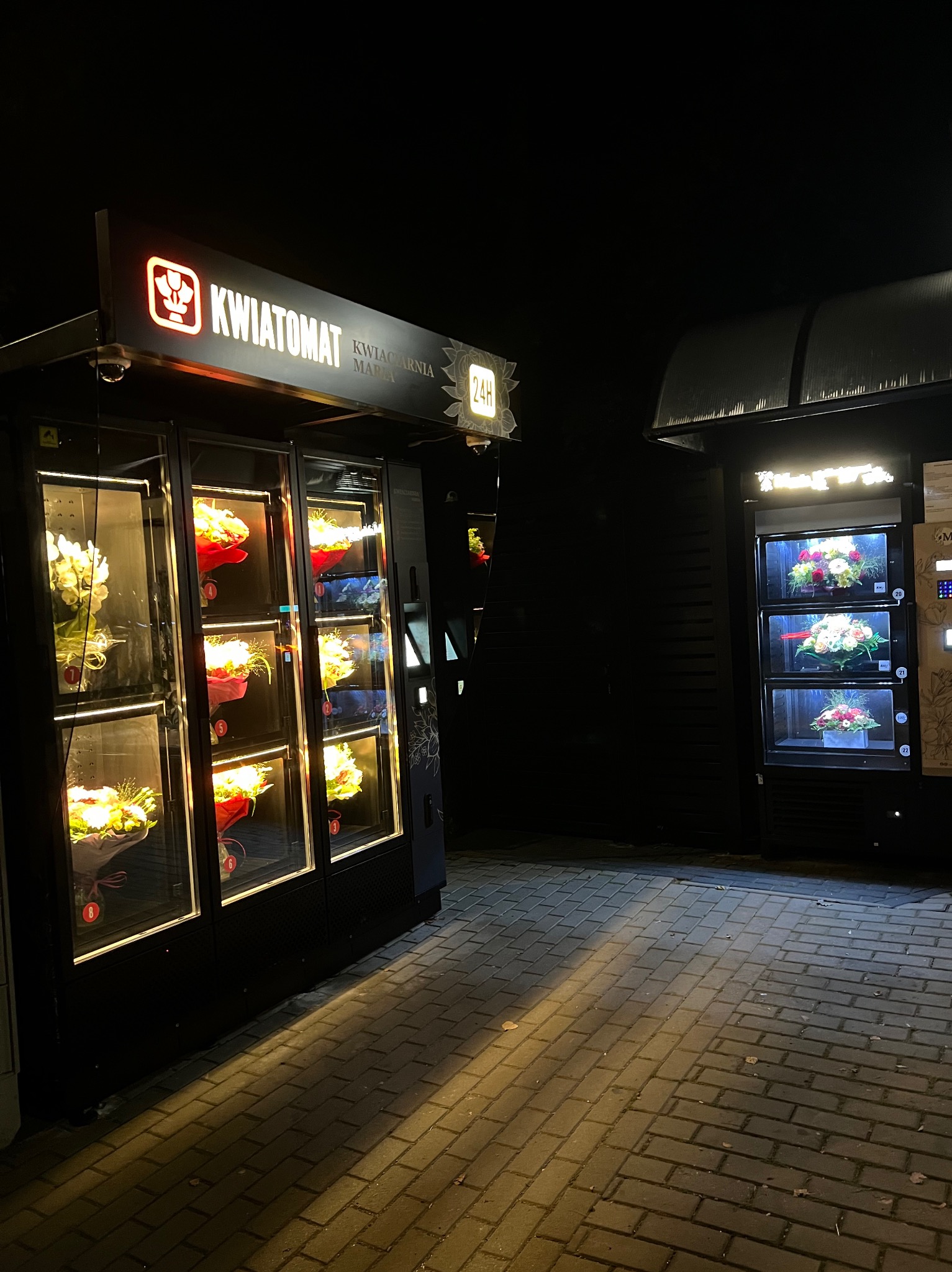 Dwa automaty kwiatowe, jeden z napisem 'KWIATOMAT KWIACIARNIA' i symbolem kwiatka, drugi z napisem 'MAJA', oba oświetlone, prezentujące bukiety w podświetlanych przegródkach, stojące obok siebie...