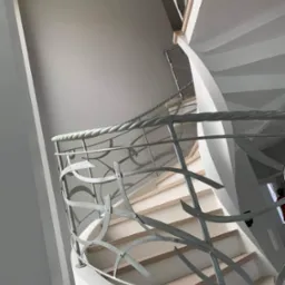 Klatka schodowa z kręconymi schodami i ozdobną, metalową balustradą w odcieniach szarości, wykończona gładzią w jasnych barwach.